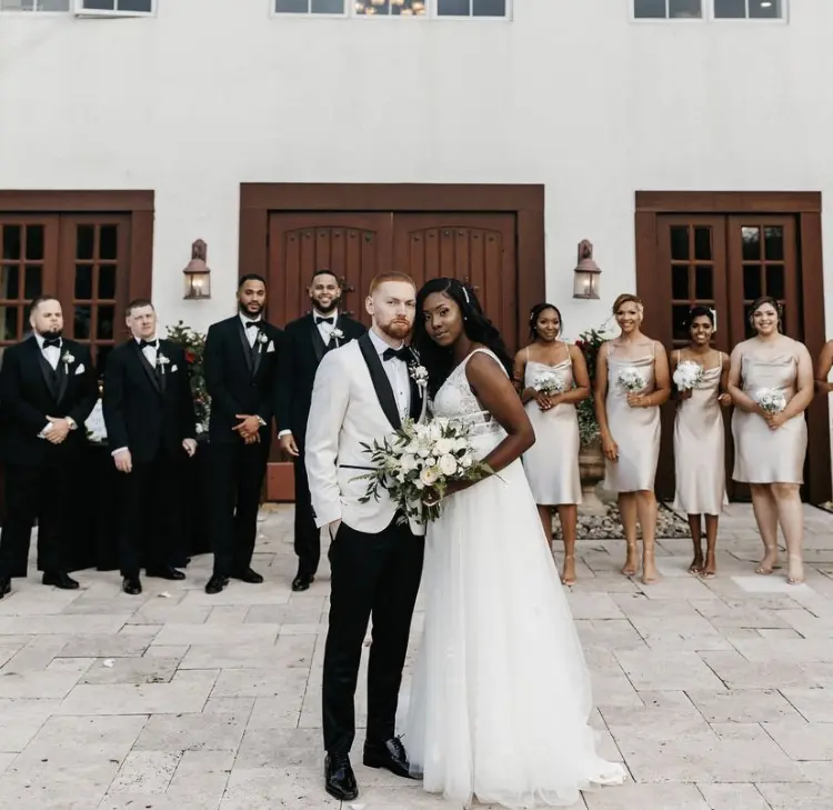 Bride wearing Raelynn wedding dress by Rebecca Ingram standing in front of her bridal party with her husband.