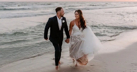 Bride wearing Lettie boho wedding dress by Rebecca Ingram holding hands on the beach with her husband.