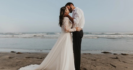 Bride wearing casual wedding dress Lorraine Dawn by Rebecca Ingram kissing her husband on the beach.