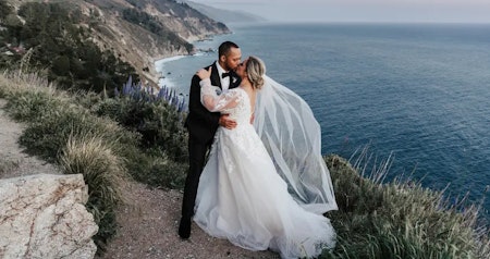 Bride wearing long sleeve wedding dress Alexandria by Rebecca Ingram on a mountain kissing her husband.